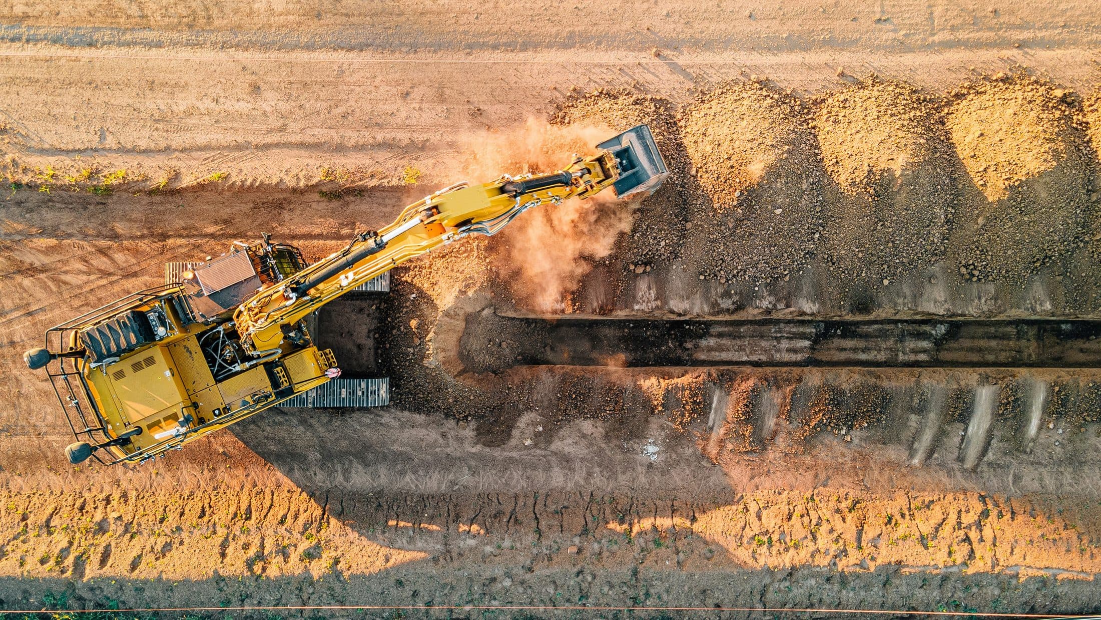 Aerial view of an excavator cutting a long drainage trench alongside a graded corridor.